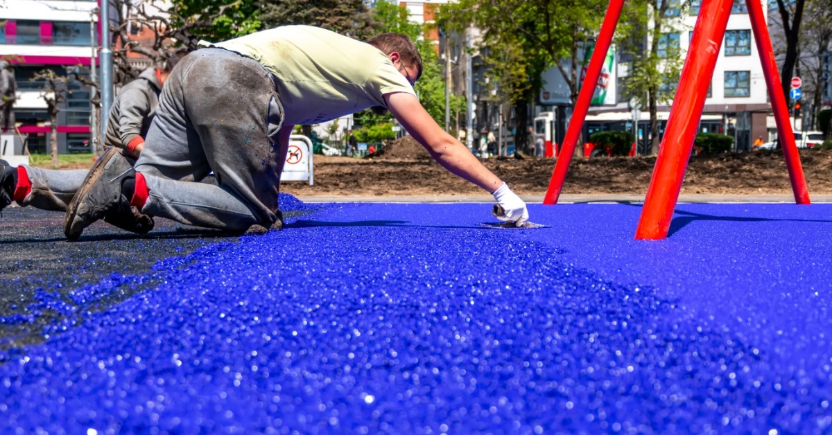 A playground professional works on their hands and knees to repair the blue portion of the playground surface.