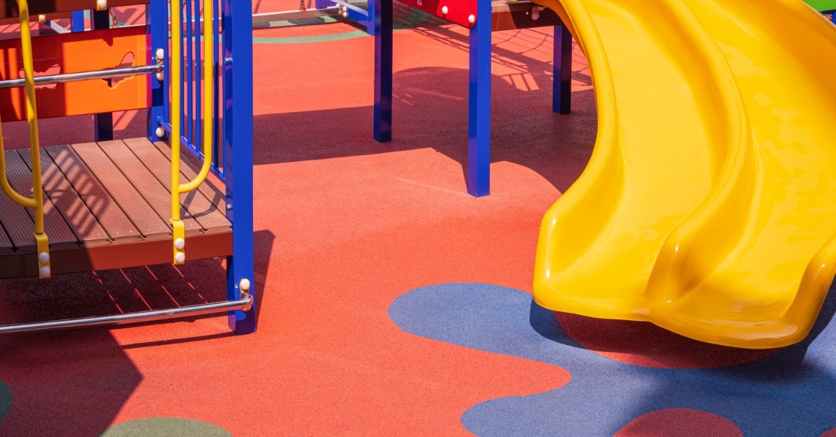 A yellow slide leads to a red and blue playground surface made of recycled rubber. Climbing equipment is on the left.