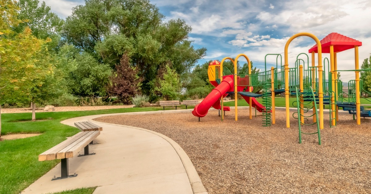 A yellow, green, and red playground set on a mulched surface. The border is a sidewalk with two bench seats.