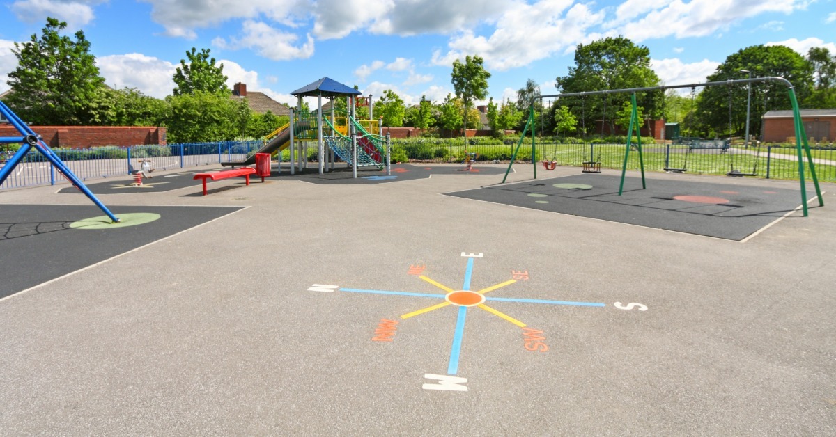 A nice, expansive school playground with a fence around the edge. There are swings, equipment, and benches.