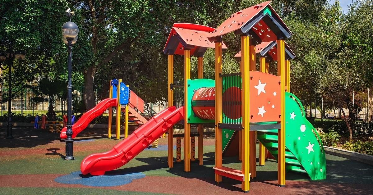 A colorful playground with slides and play structures on soft, colorful rubber flooring in a sunny park among trees. A colorful playground with slides and play structures on soft, colorful rubber flooring in a sunny park among trees.
