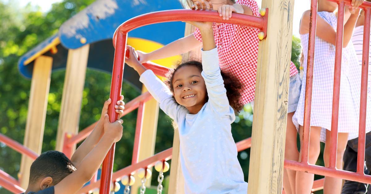 Smiling children play on a playground structure, with one girl hanging from a bar while others climb and watch.