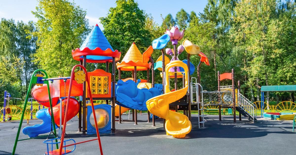 A colorful playground with slides, climbing features, and decorative towers set on a rubber surface, surrounded by green trees.