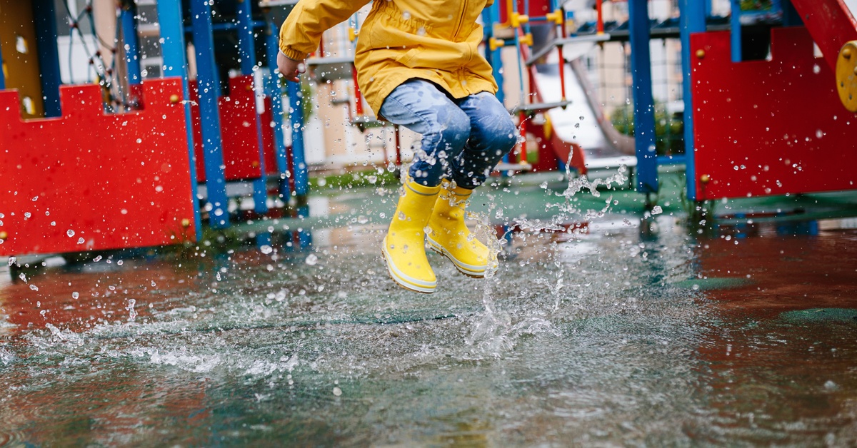 How Playground Borders Influence Drainage and Erosion A child in yellow boots and a yellow rain jacket jumps in a puddle on a flooded playground with red and blue equipment.