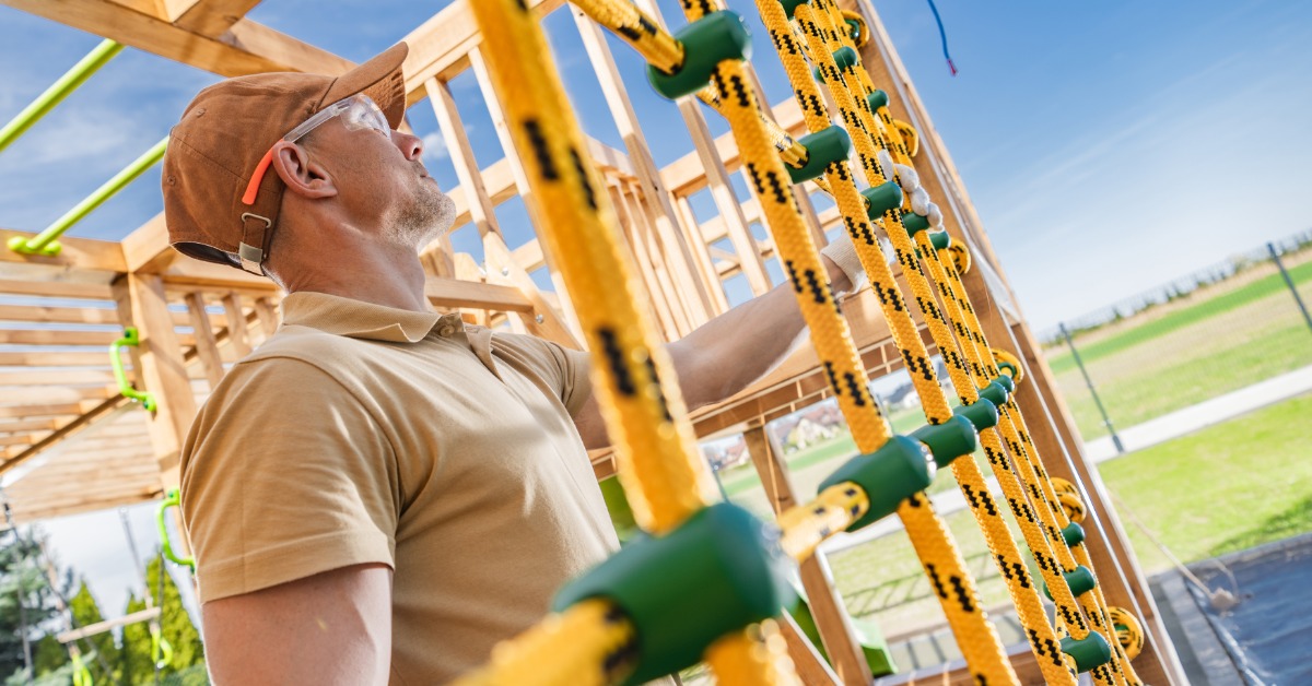 How To Budget for Playground Repairs and Upgrades A maintenance worker inspects and tightens a rope climbing net on a wooden outdoor playground structure.