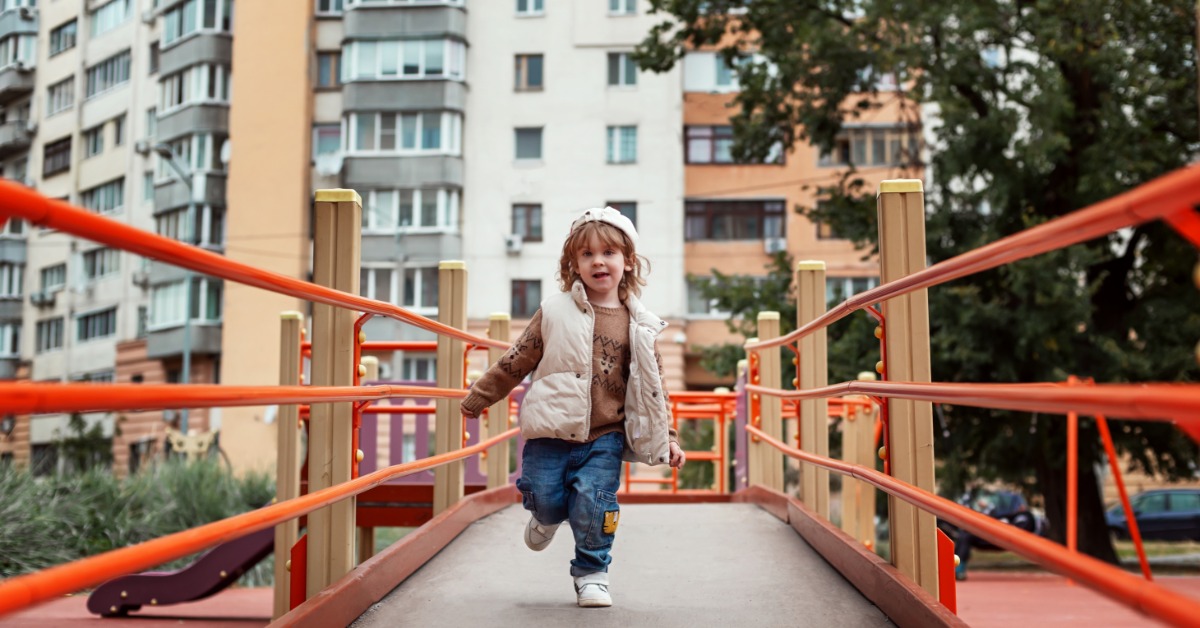 A young child walks across a playground ramp with bright orange railings in a residential park setting.