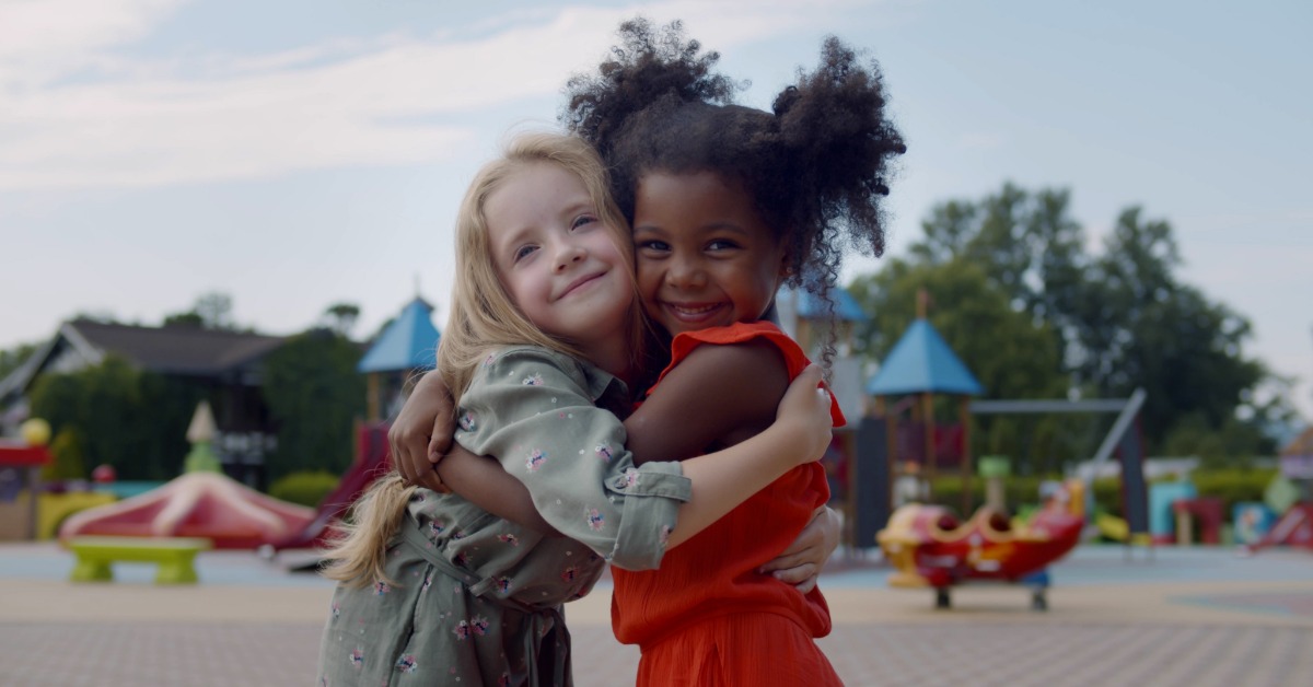 Two young girls hugging and smiling at a colorful daycare playground, with rides and play structures in the background.