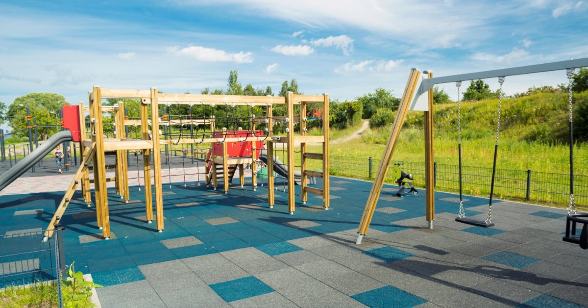 An outdoor playground with a wooden climbing structure, monkey bars, swings, and rubber tile safety surfacing on a sunny day.