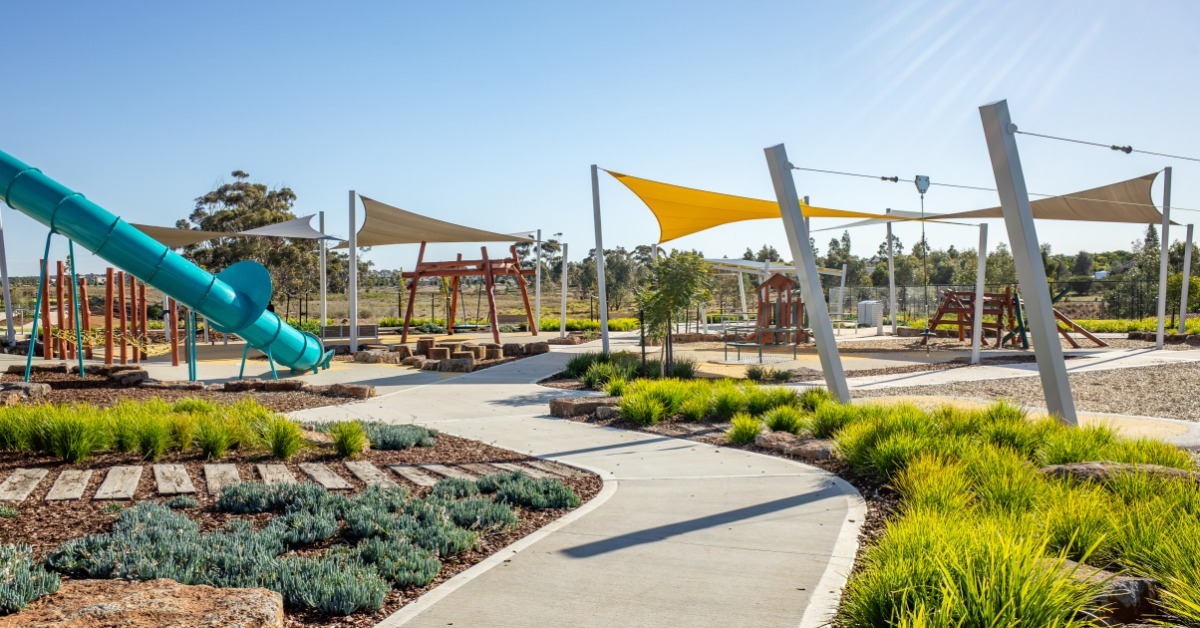 An open playground area with yellow and white shade sails, a teal tube slide, and winding paved paths through green landscaping.