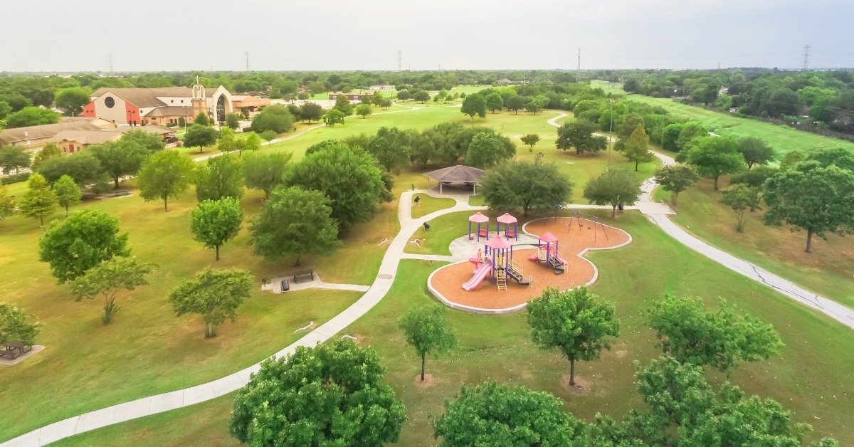 An aerial view of a community park with a colorful playground, pavilion, walking paths, and tree-lined green space.